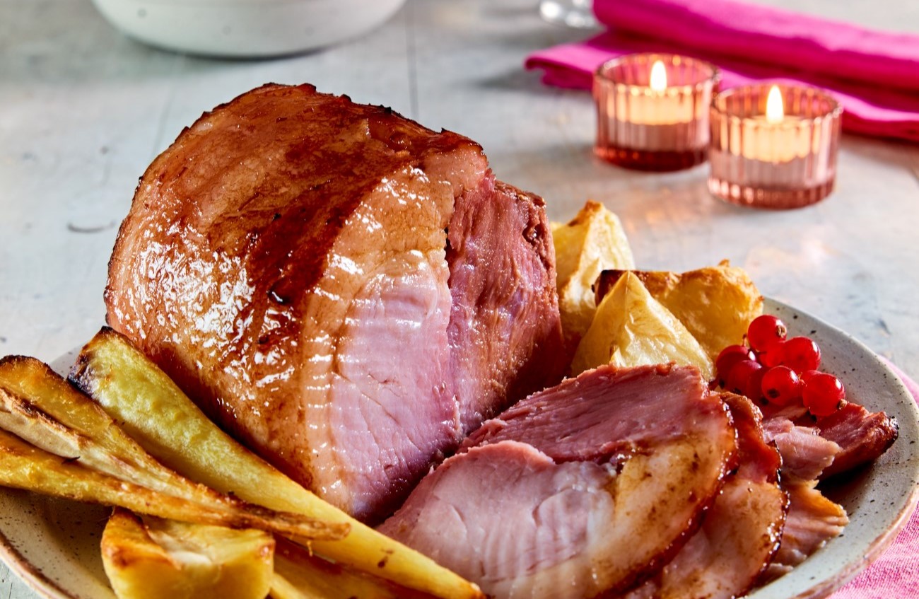 Roast gammon joint, some sliced, on a serving plate, with vegetables.