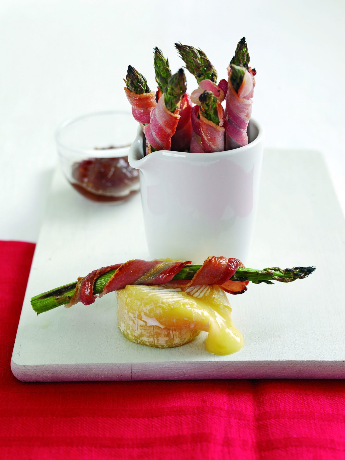 Bacon wrapped asparagus in a white pot, on a white plate. Gooey cheese shown on plate.