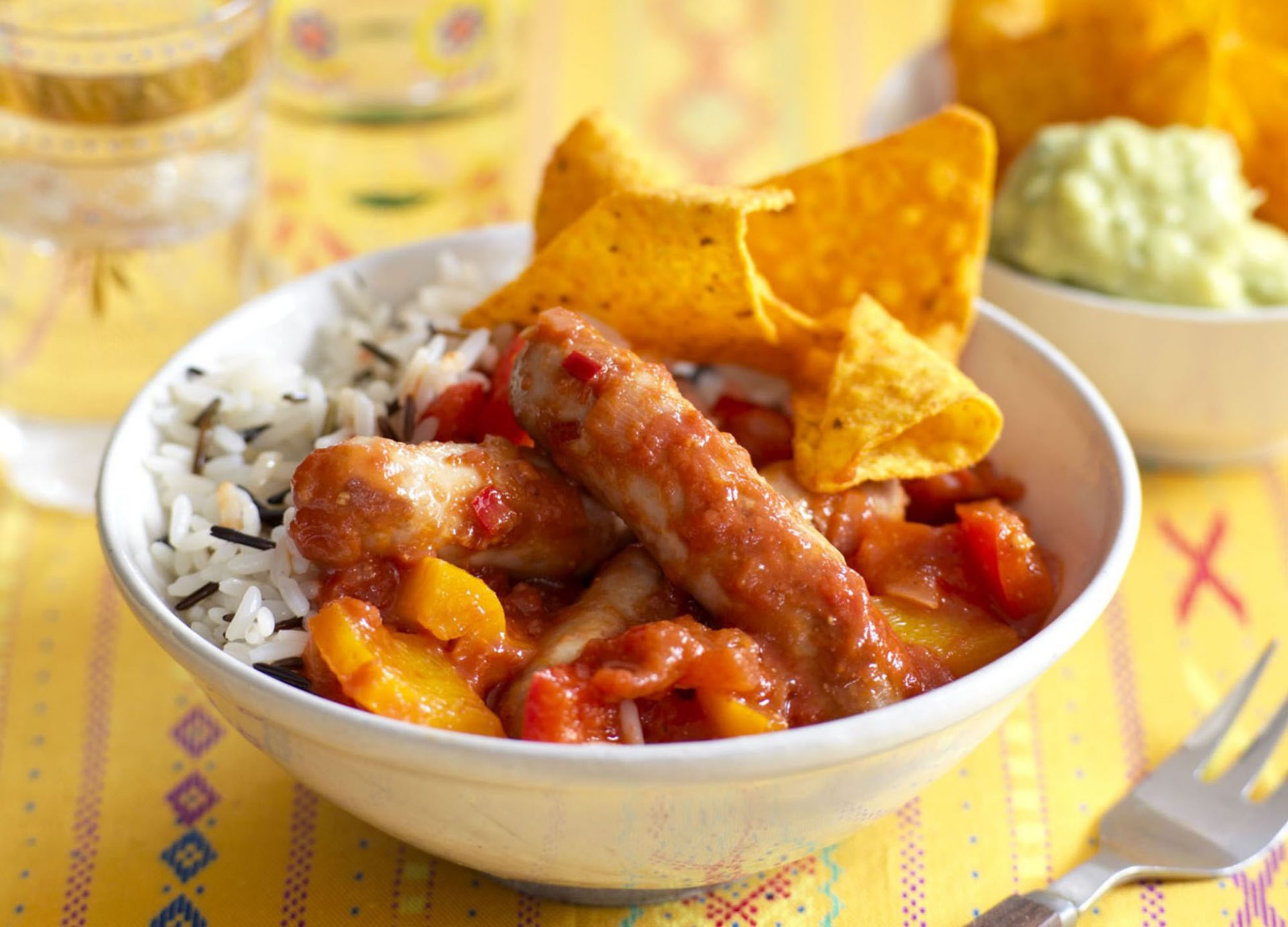 Chipolata sausages with chilli and rice with tortilla crisps, in a white bowl.