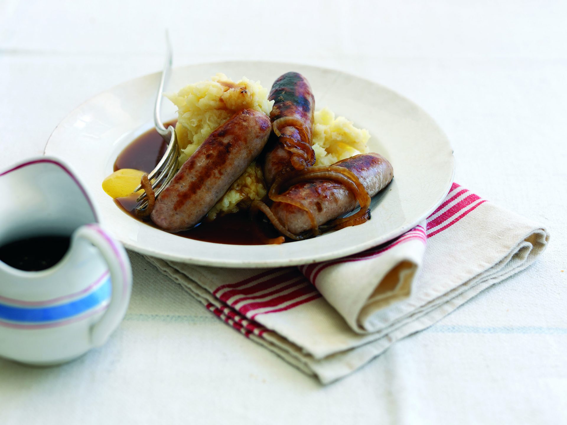 Three pork sausages on a bed of rooty mash, on a white plate. Small jug on the side.