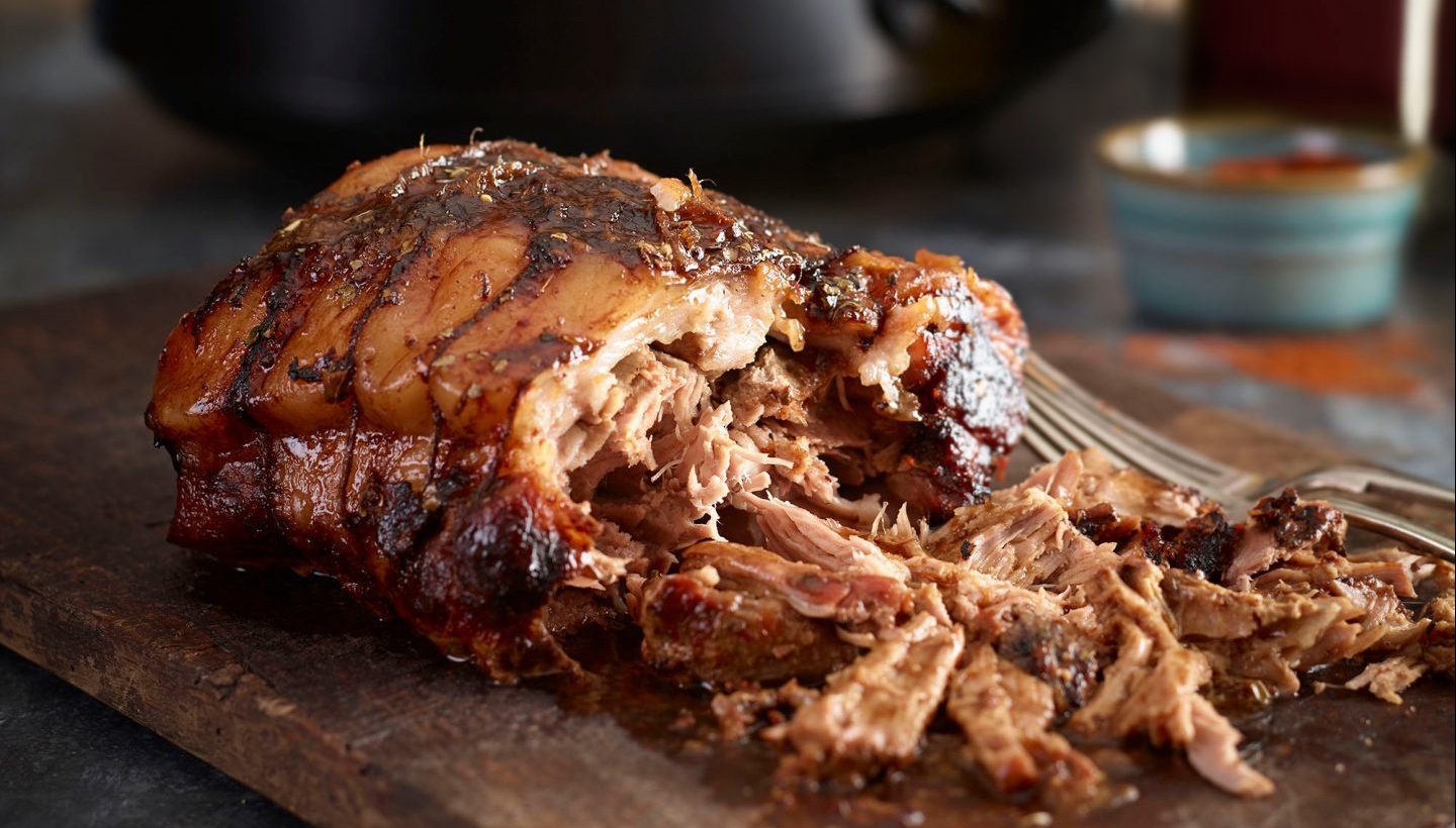 Part shredded pulled pork on a wooden board, with slow cooker in background.