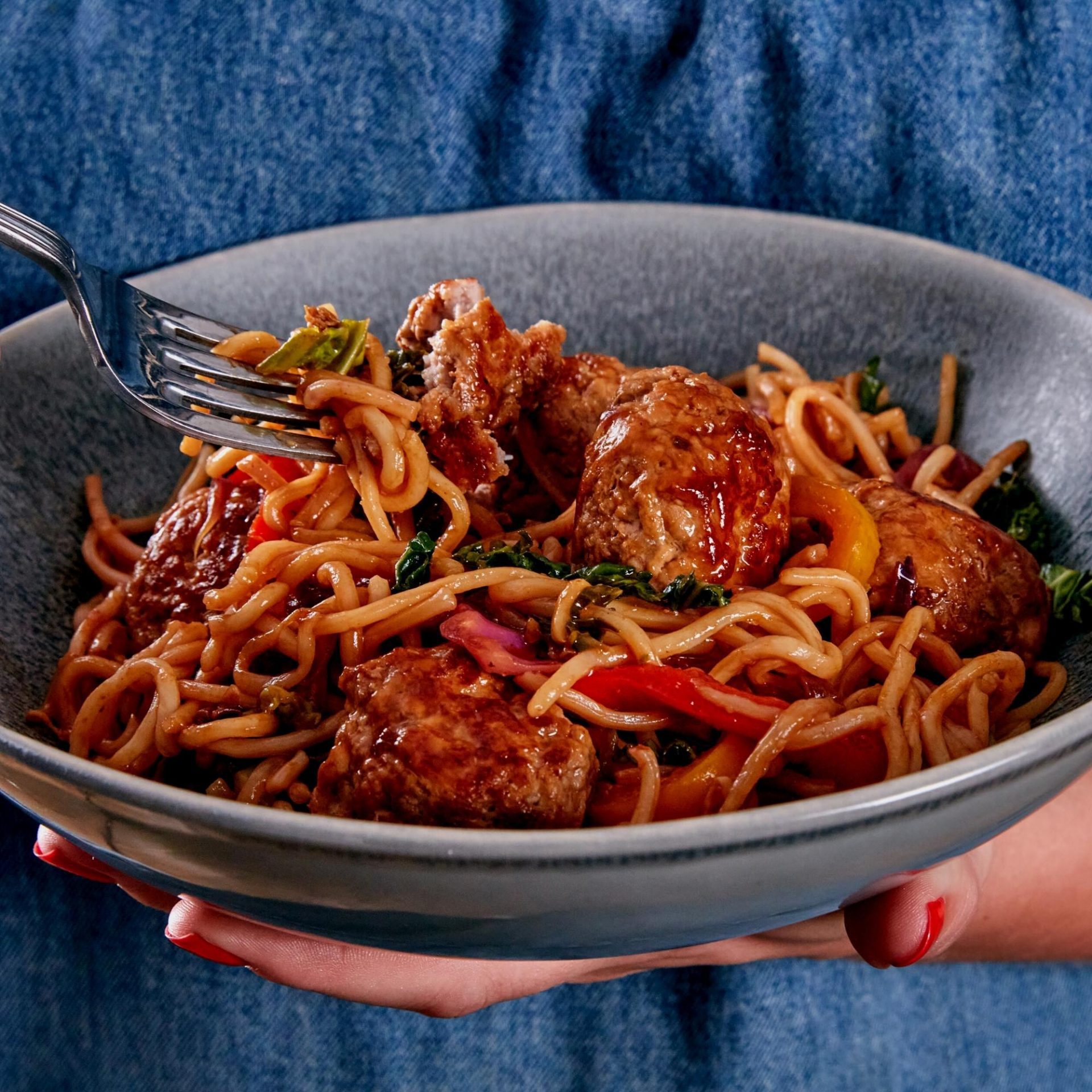 Hand holding a bowl containing stir fried pork meatballs and noodles, fork in shot