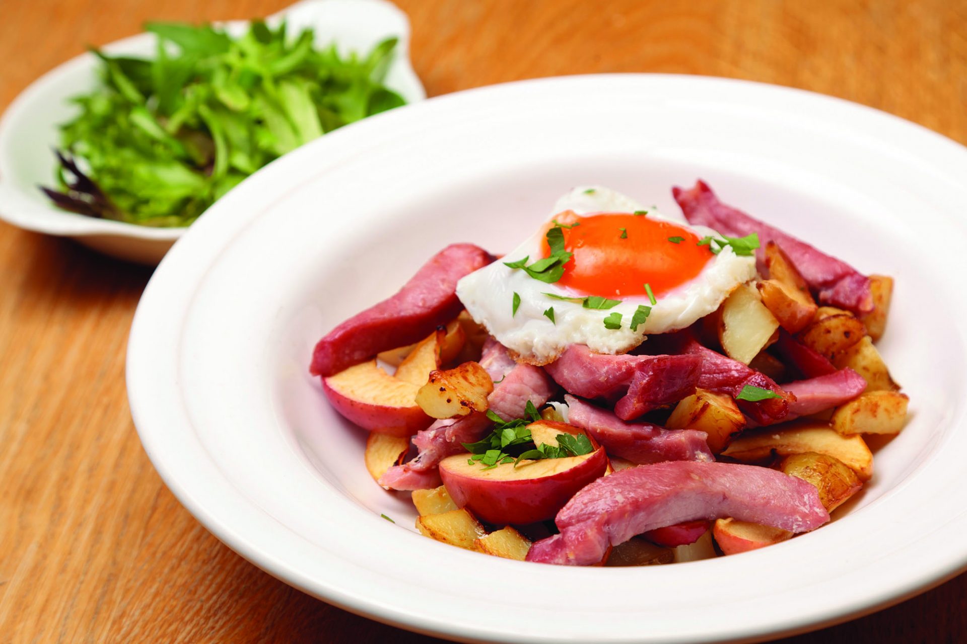 Gammon potatoes and apples in a white dish with fried egg on top.