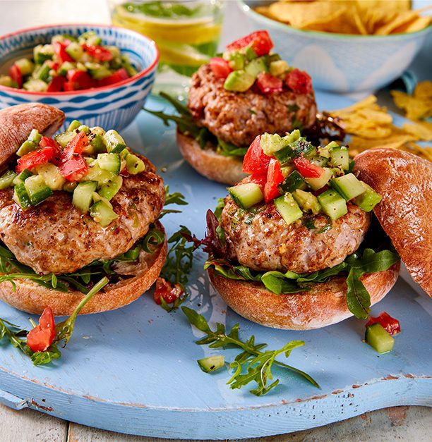 Three burger buns filled with pork burger and an avocado and tomato salsa, on a pale blue board.