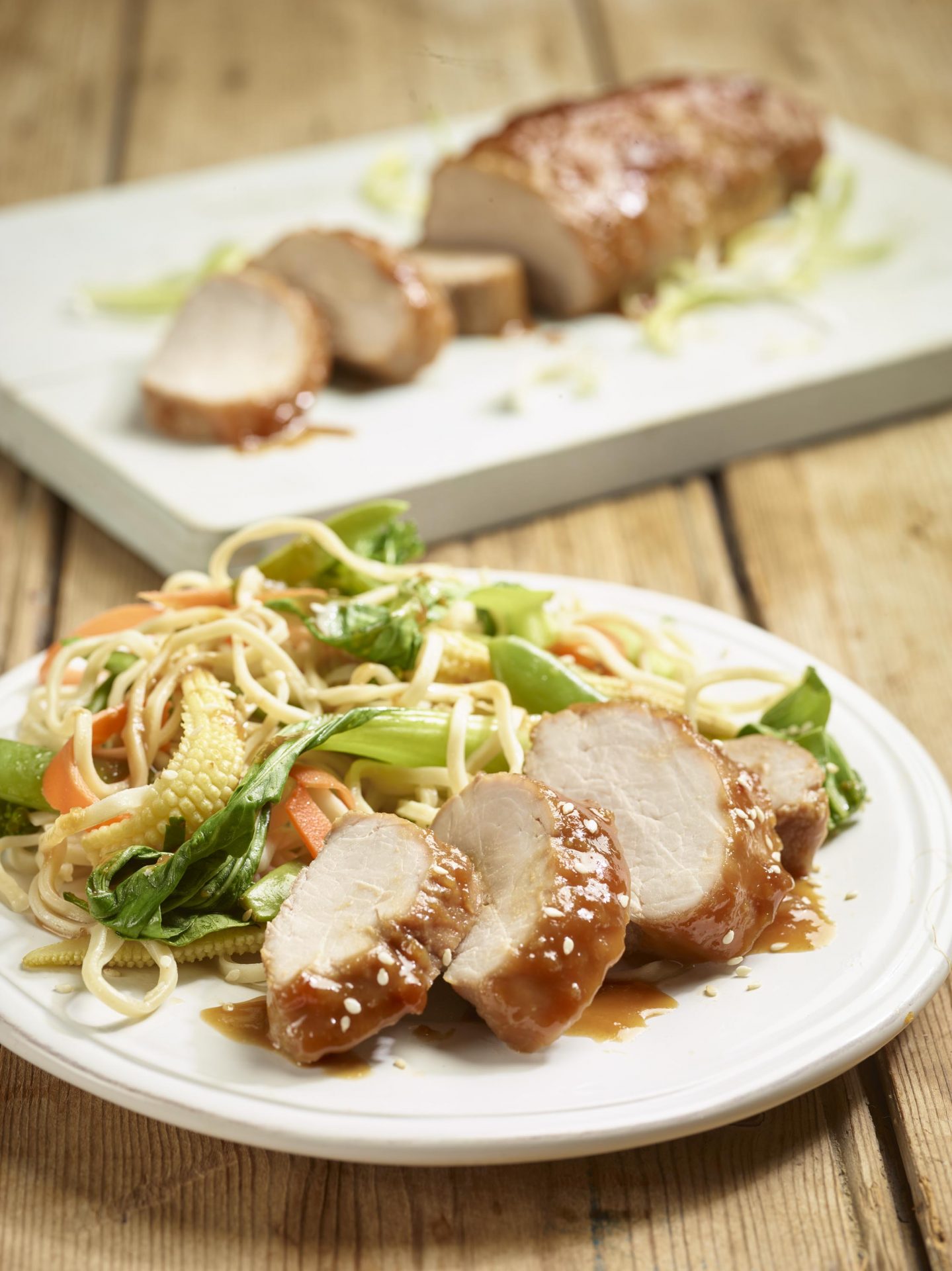 Sliced miso pork fillet served with vegetables and noodles on a white plate. Pork fillet in background