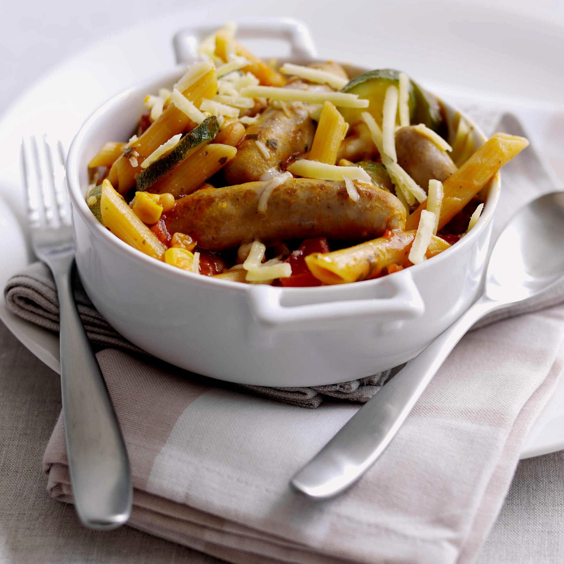 Chipolata sausage bean pasta in a white casserole dish, with cutlery on side.