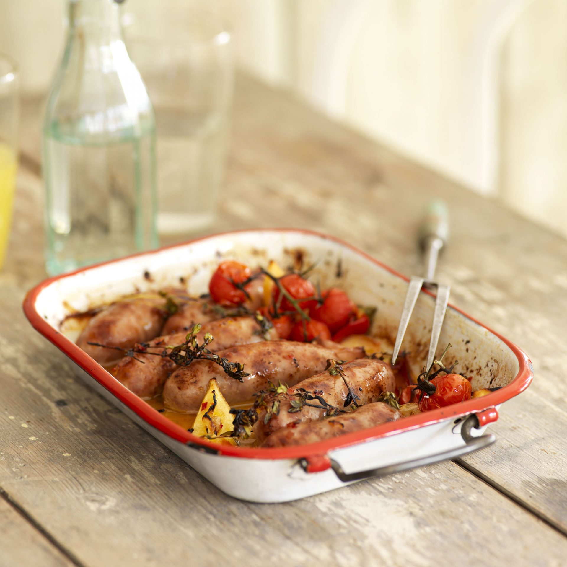 White baking dish containing piri piri style baked sausages and vegetables, on a wooden table