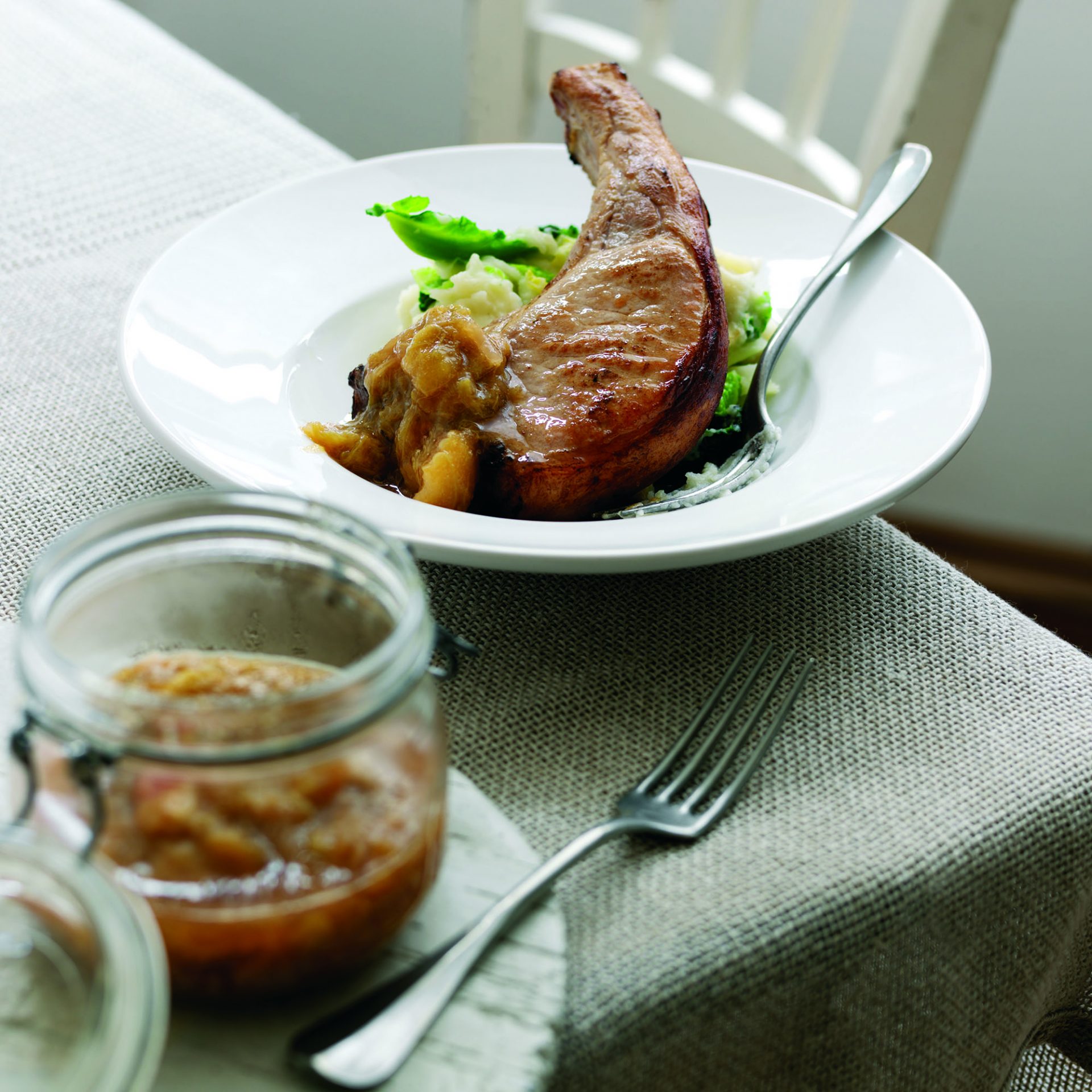 Pork chop with rhubarb apple and ginger chutney served in a white dish, with chutney jar on side.