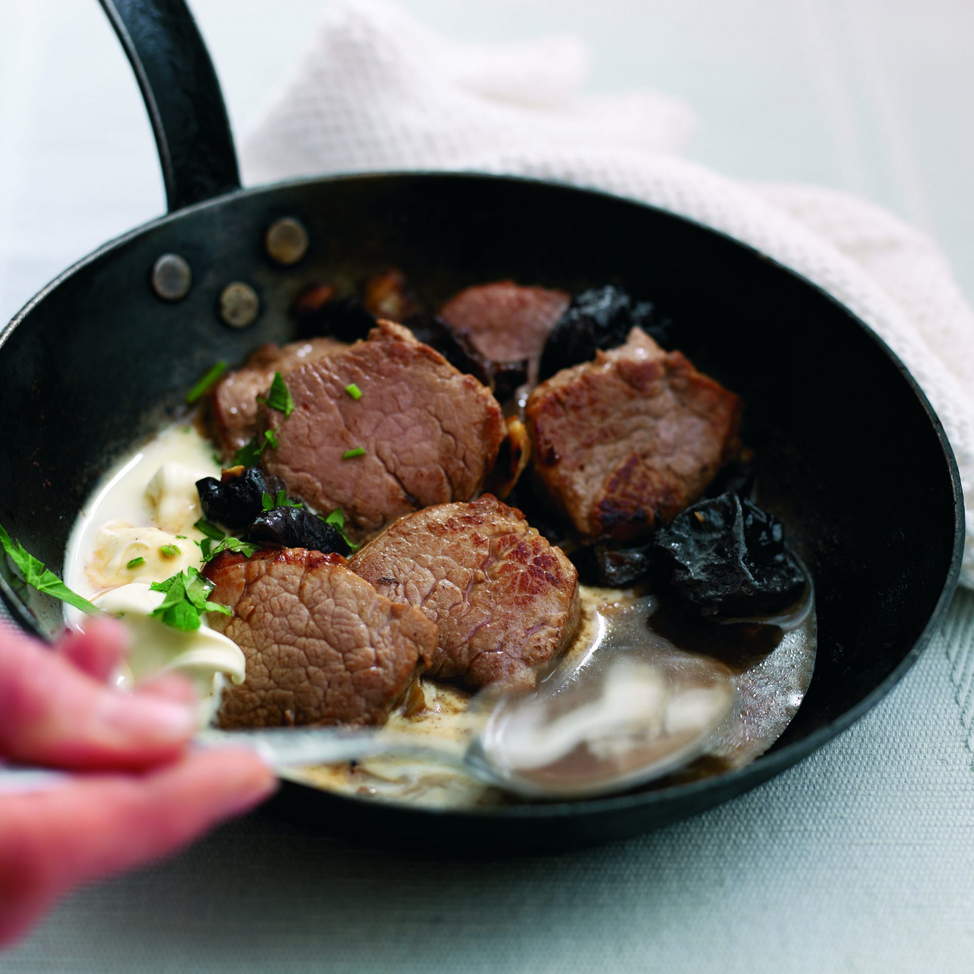 Pork fillet with creamy wine and prune sauce in a black frying pan with spoon scooping sauce