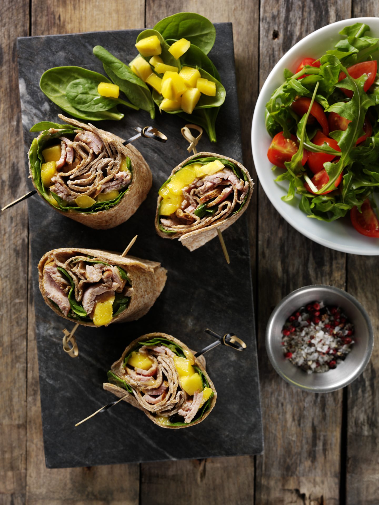 Four spinach mango and pork wraps on a black board, with side salad