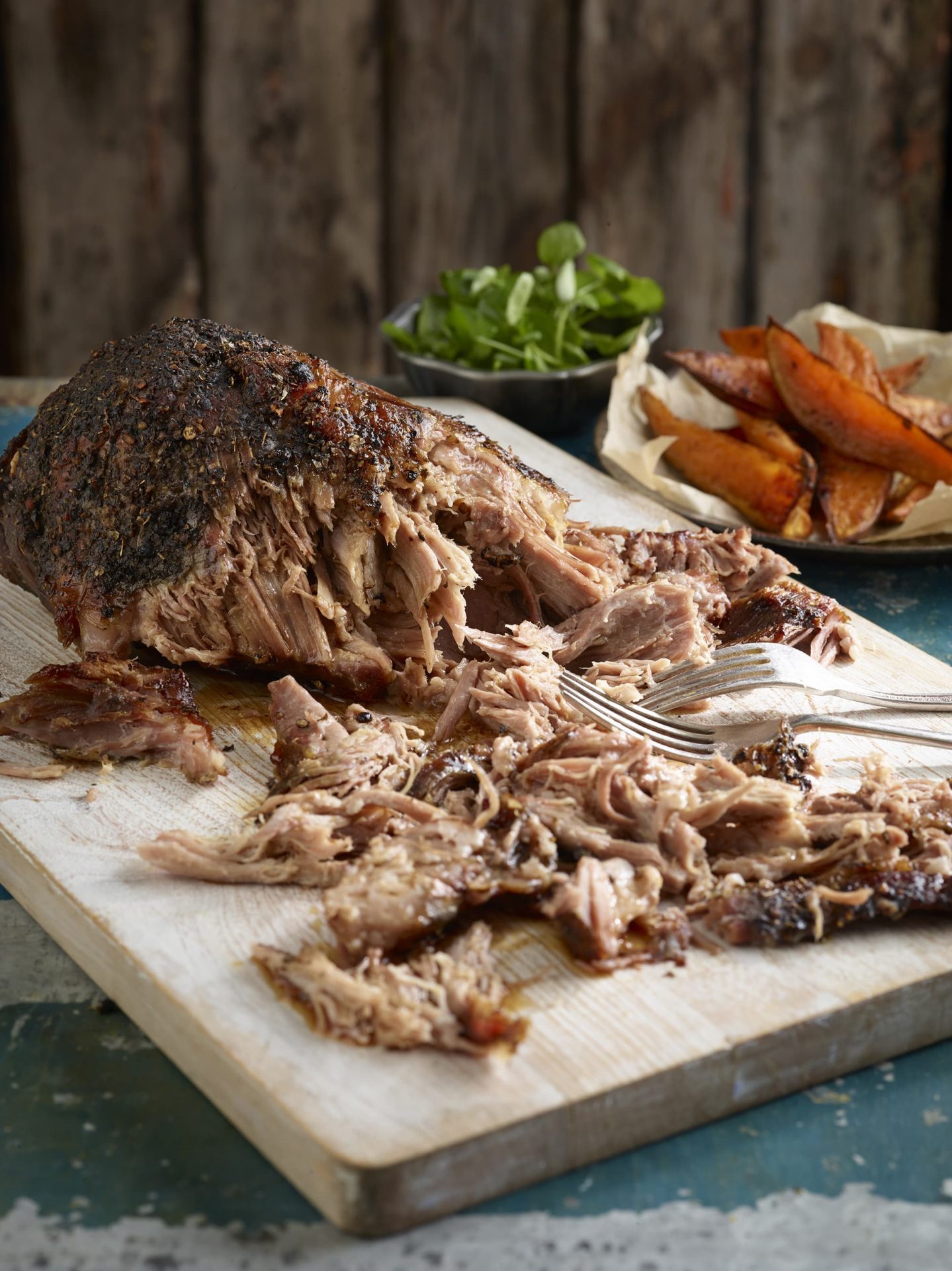 Pulled pork joint, some pulled, on a wooden board with two forks for pulling