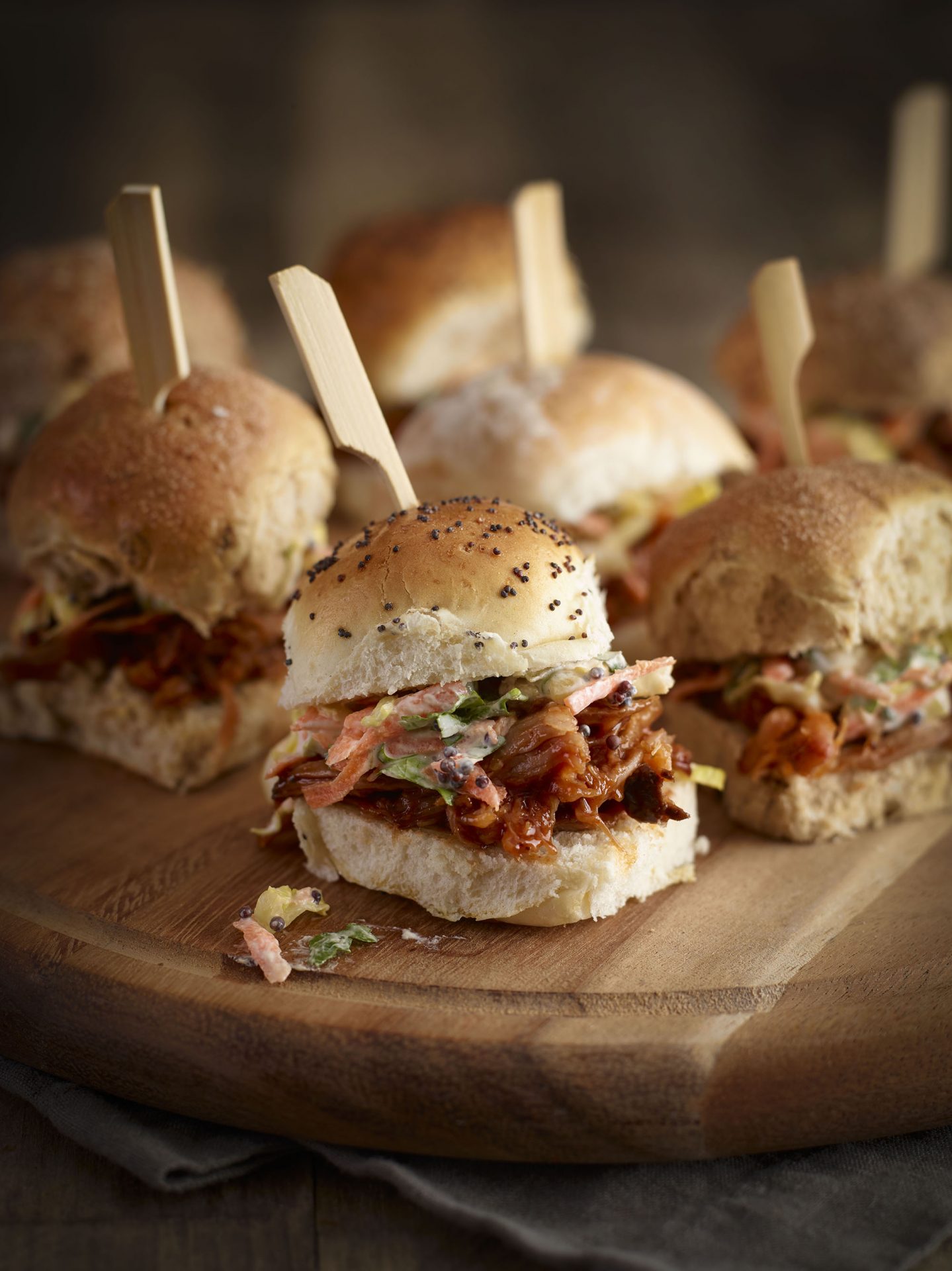 Small burger buns filled with pulled pork and coleslaw with a skewer in the top, served on a wooden board