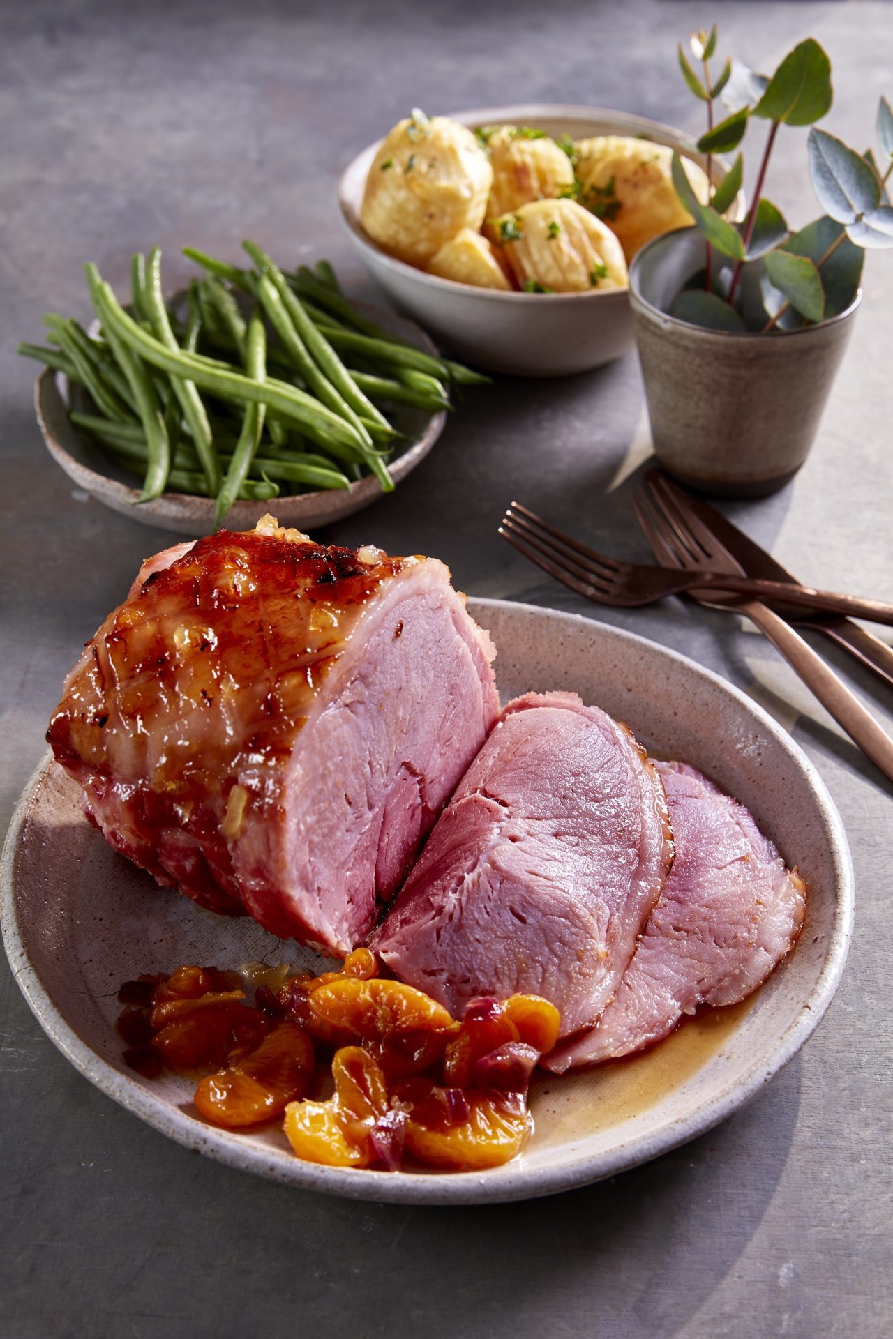 Roast gammon joint, some sliced, on a white plate with green beans and vegetables in background