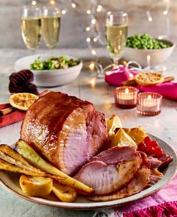 Roast gammon joint, some sliced, on a serving plate, with vegetables. Candle lights and champagne glasses in background