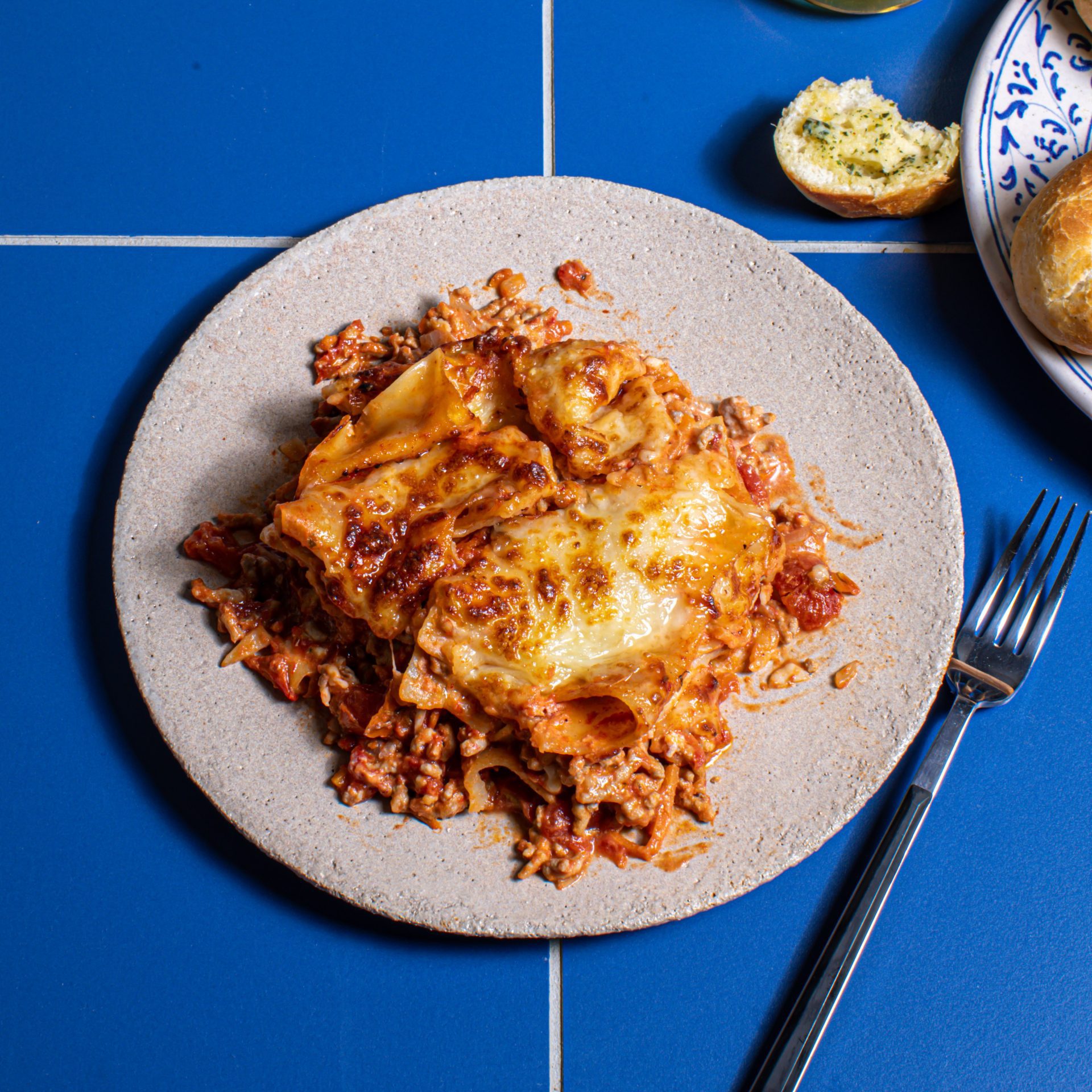 Pork mince lasagne on a pale plate with fork on a blue background.