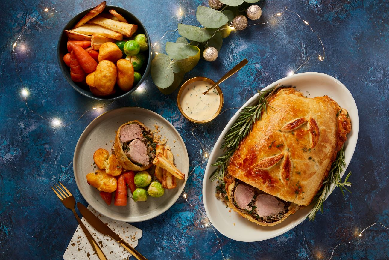 A selection of dishes on a festive themed dinner table showing a whole Pork Wellington, a serving of Wellington with vegetables and then a bowl full of potatoes, parsnips, carrots and Brussel sprouts