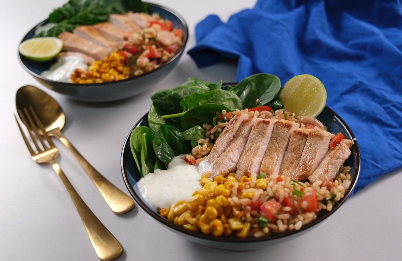 A pork steak protein bowl in a bowl with gold cutlery and blue cloth.